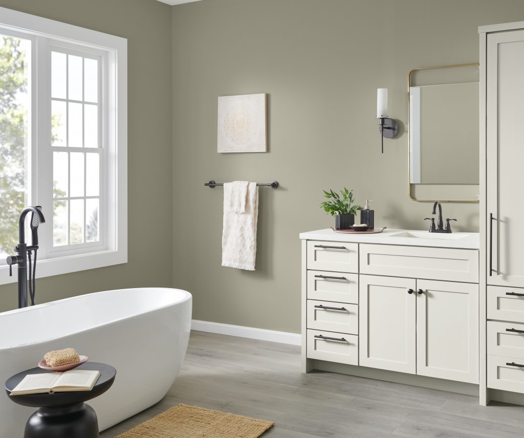 A spacious bathroom with walls painted in a grey-green colour, styled with a freestanding tub and a white vanity and cabinets
