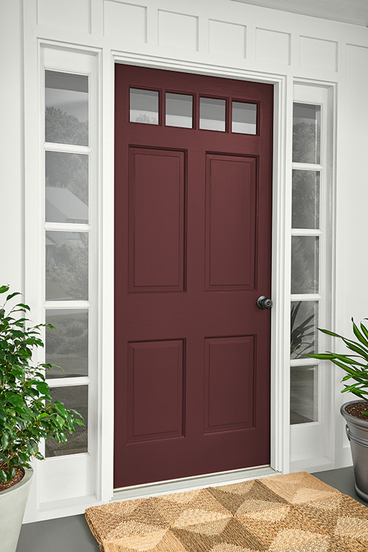 A red front door set within a white door frame. The door has a silver doorknob on the right side. To the left of the door, there’s a part of a houseplant visible, and in front of it is a beige and brown patterned doormat on grey flooring. 