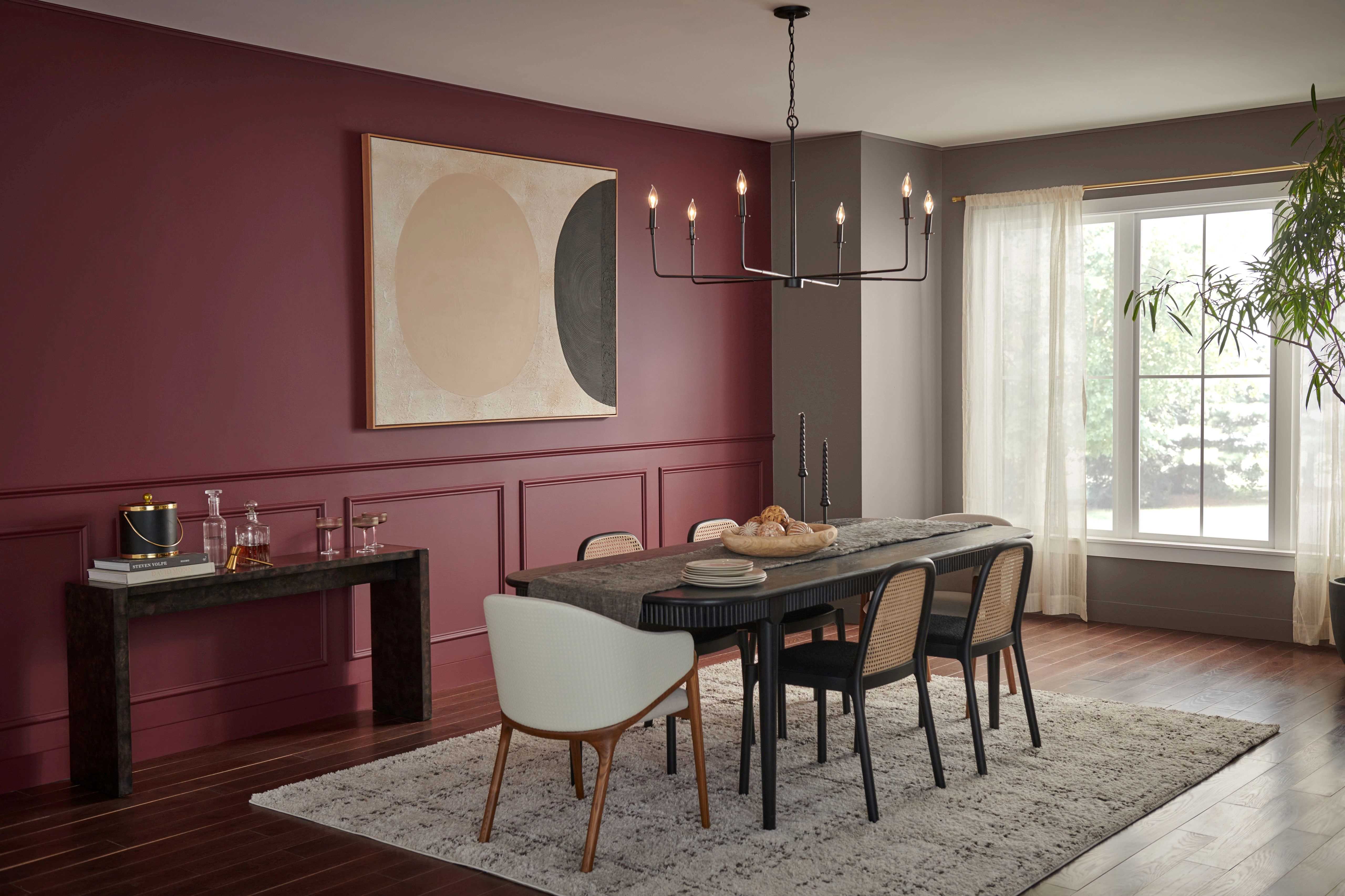 An elegant dining room featuring a large rectangular table with a dark finish, surrounded by six modern chairs. Above the table hangs a sophisticated chandelier with multiple arms and candle-like lights. The room is defined by a deep red accent wall with wainscoting details, this wall hangs an abstract painting with beige, black, and white circles.  