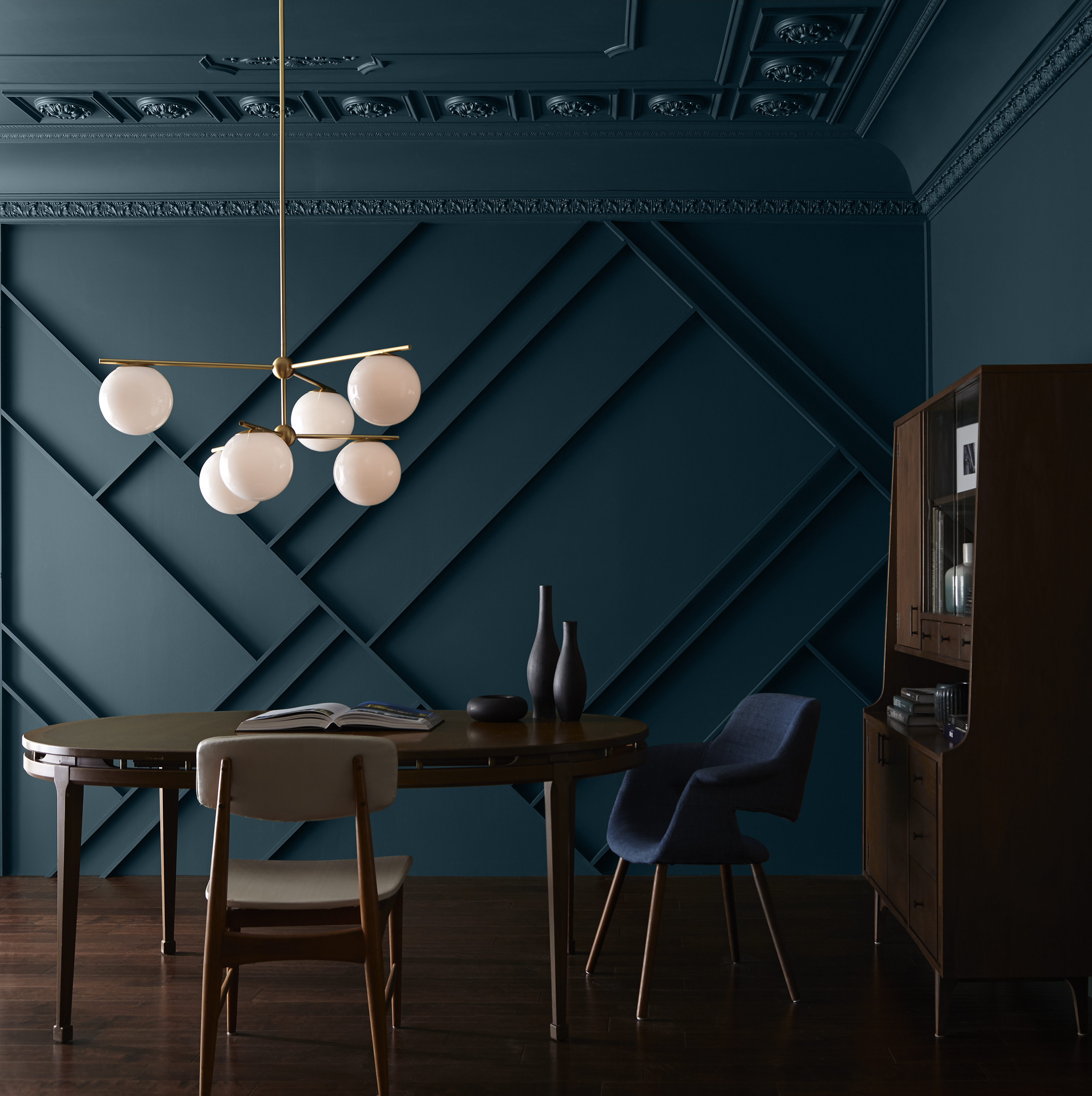 An elegant dining room with dark teal-blue walls adorned with geometric panelling. A modern chandelier hangs above a wooden table, accompanied by matching chairs and a bench. To the right, a wooden cabinet displays decorative vases, enhancing the room’s sophisticated aesthetic.
