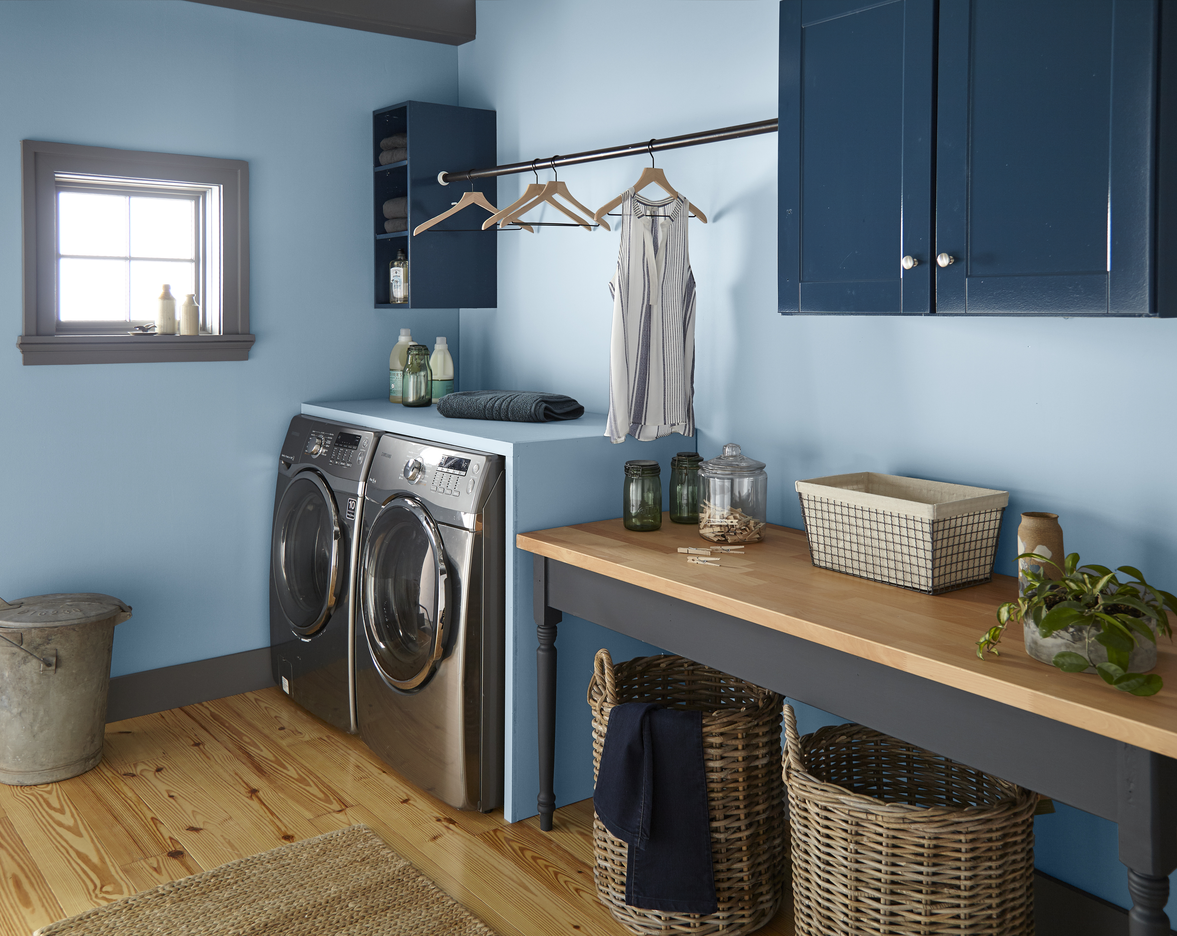 A laundry room showcasing storage solutions for laundry essentials and clothes hanging space. The walls are painted in a midtone blue shade, and there are closed upper cabinets painted in a deep blue colour called Inked. 
