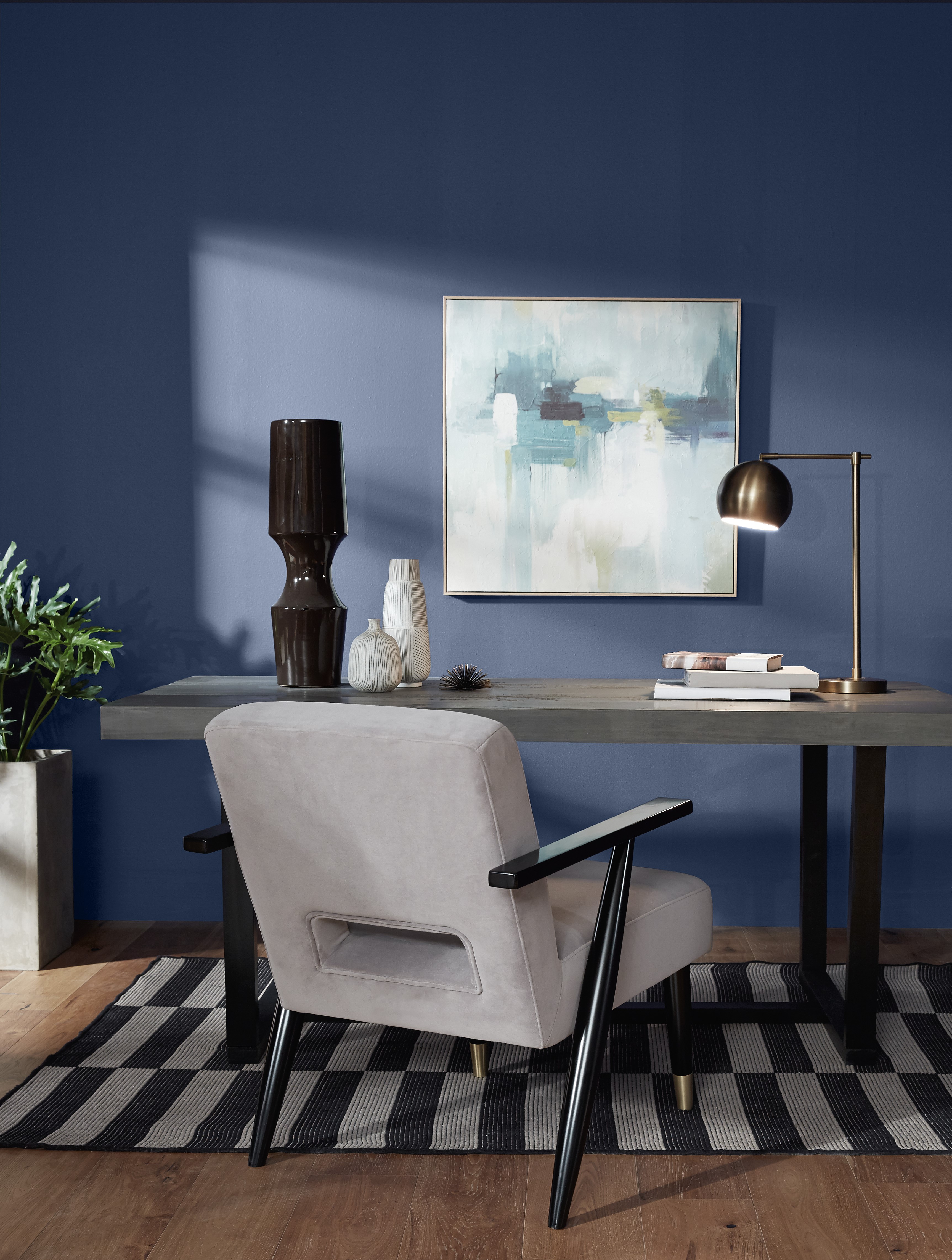 A modern home office interior featuring a dark wooden desk with a beige upholstered chair with black armrests. On the desk, there’s a black lamp with a gold interior shade and two closed books stacked beside it. Above the desk hangs an abstract painting with blue, white, and beige tones in a gold frame. The room has dark blue walls and is lit by natural light casting shadows on the wall and patterned black and white rug beneath the furniture.
