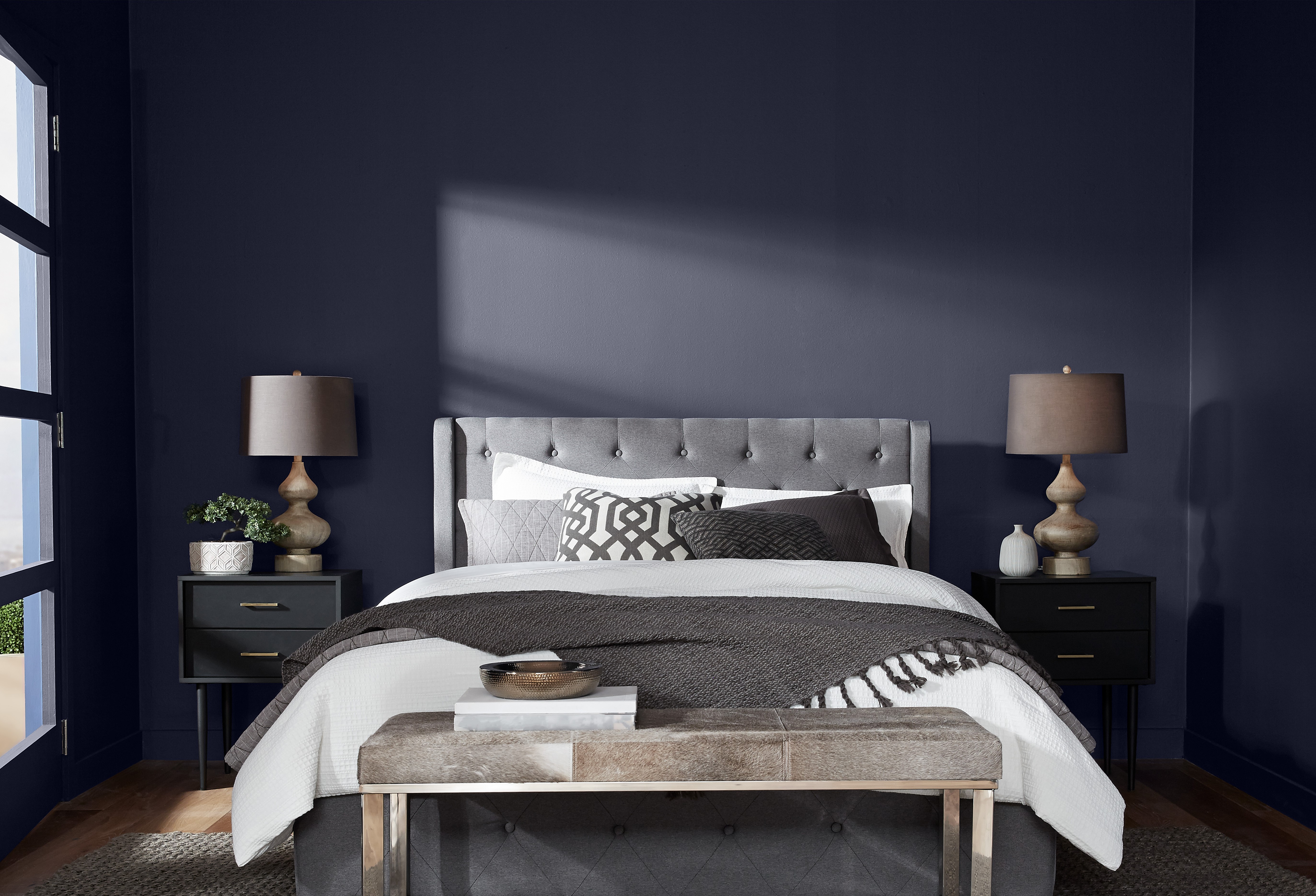 A bedroom image that shows dark blue painted walls.  The room is well-organized with a large bed featuring a grey tufted headboard centered against a deep blue wall. The bed is adorned with white, grey, and patterned bedding, along with multiple pillows. The room has dark hardwood floors and is illuminated by natural light from a window on the left, partially covered by drawn curtains.
