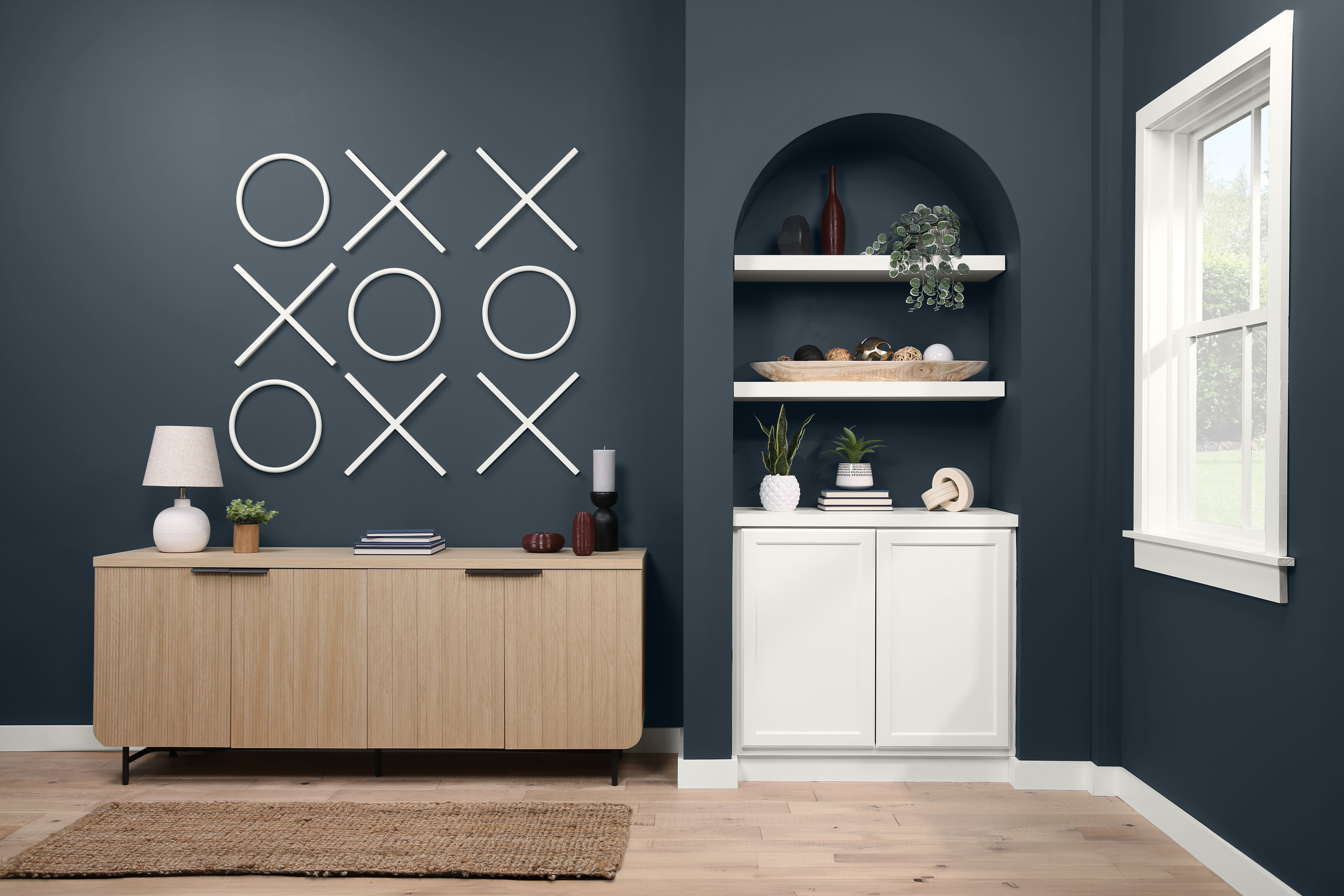 The interior of a stylish home office with playful décor featuring a large tic-tac-toe  game as wall art. A modern sideboard is adorned with a white lamp and a built-in shelving unit with an array of decorative items and greenery.  The dark blue room has a window that allows planty of light to come through, enhancing the serene and modern atmosphere. 