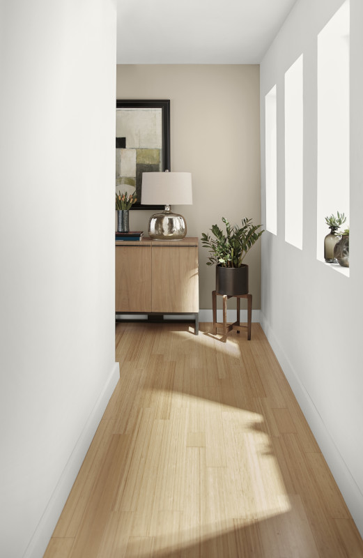 A long hallway with pristine white walls that reflect the ample natural light streaming in from large windows. The golden tone from the floor enhances the brightness of the space.  At the far end, a piece of modern art in vibrant colours contrasts beautifully with the white and  beige tone surroundings, adding a touch of personality to the hallway.