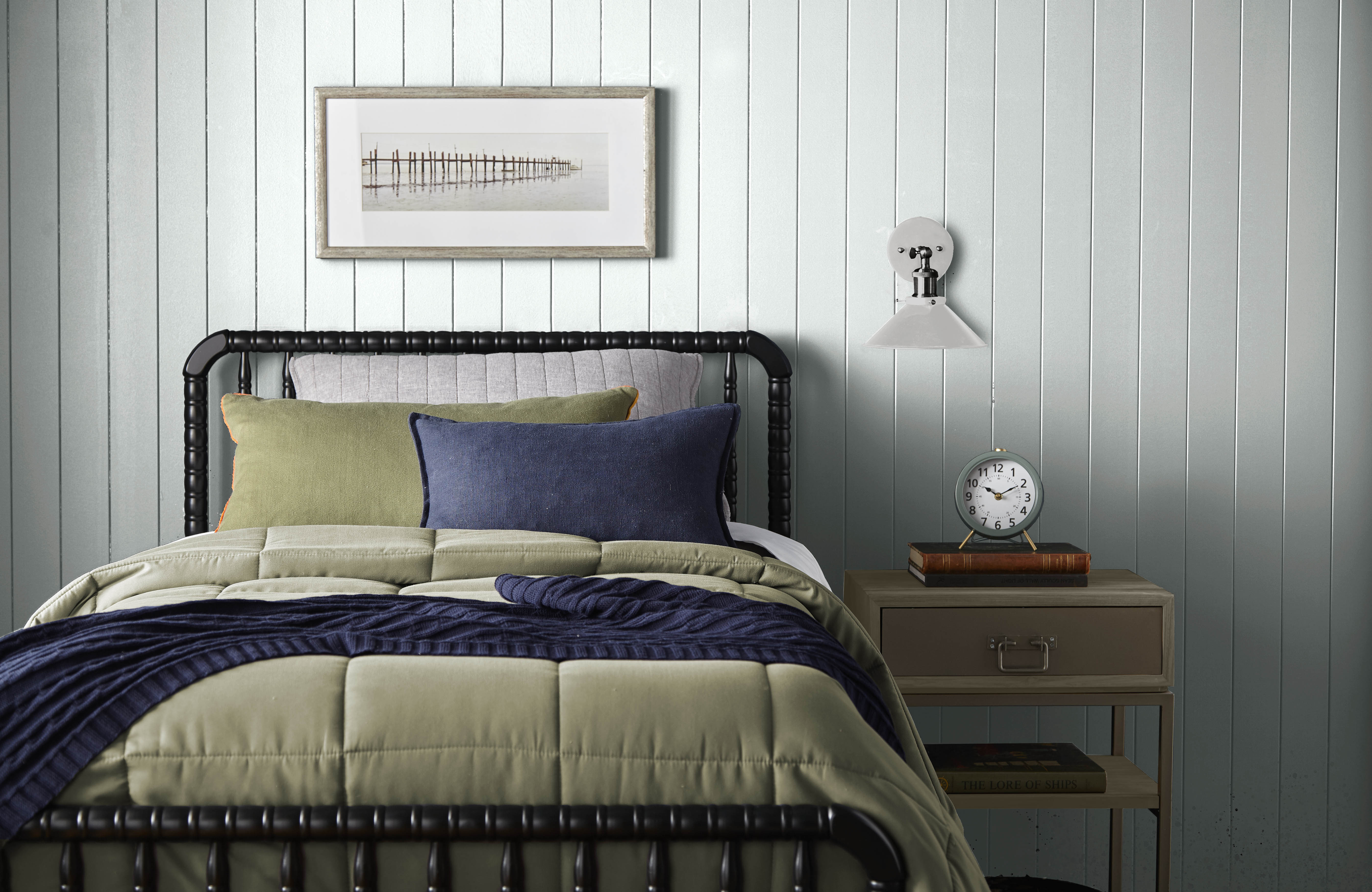 A bedroom featuring a soothing bluish white wall with vertical beadboard panelling that adds texture and depth. The room is furnished with a black metal bed frame holding neatly arranged pillows in shades of green and blue, complemented by a quilted comforter in navy blue and beige. 