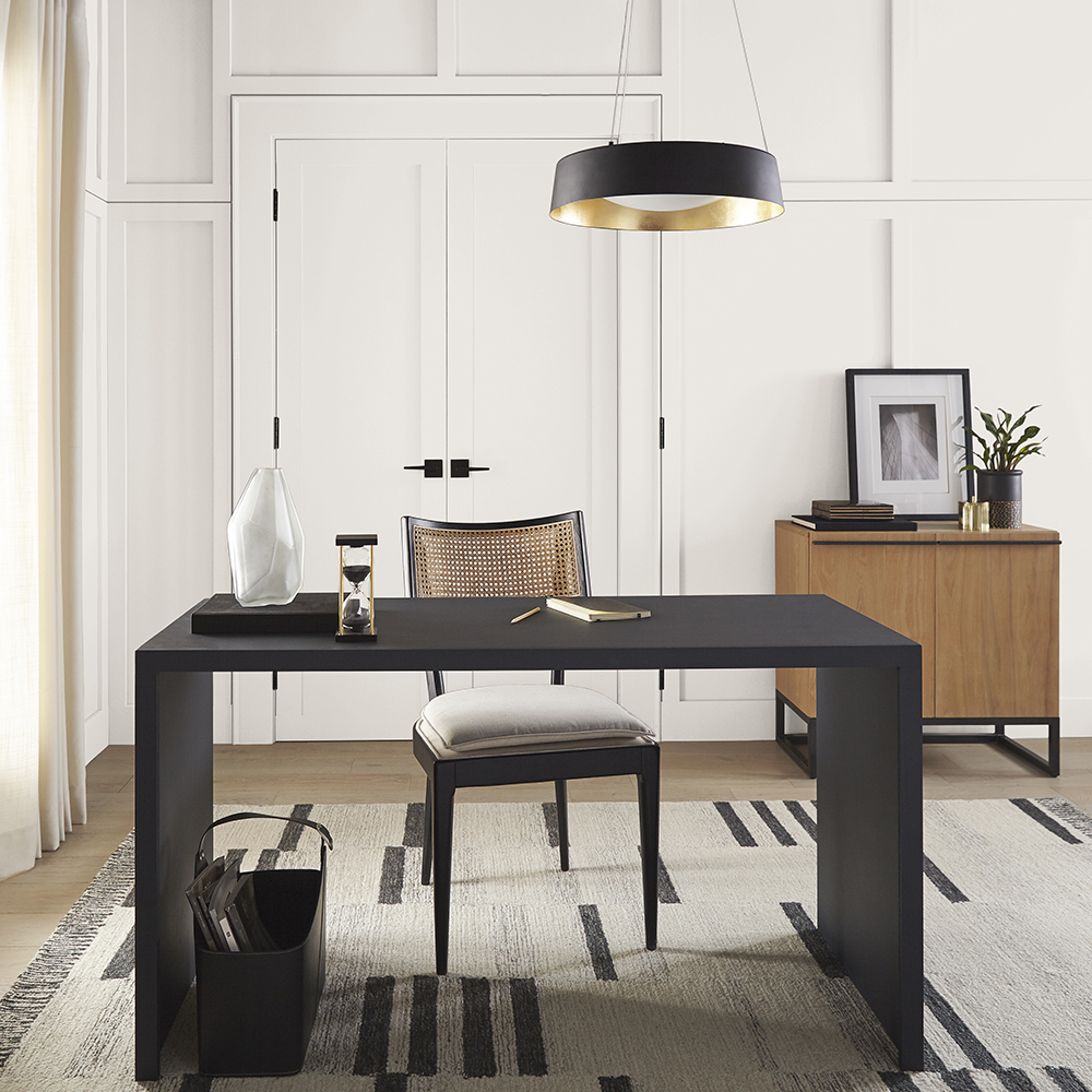 A contemporary home office featuring Silky White walls. At the center of the workspace is a sleek, black rectangular desk paired with a matching chair. The floor is adorned with a geometric-patterned rug in shades of black and greige.