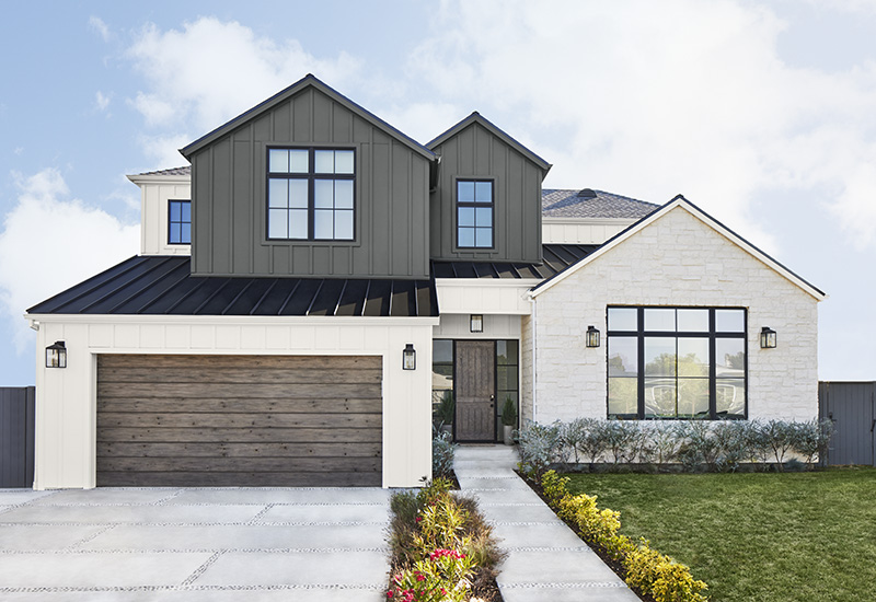 A large modern farmhouse featuring a striking white exterior with contrasting dark grey accent colour and black roofing and accents. The structure showcases a mix of textures, including white brickwork and vertical siding. The garage door is wooden with horizontal planking, adding warmth to the cool colour palette.