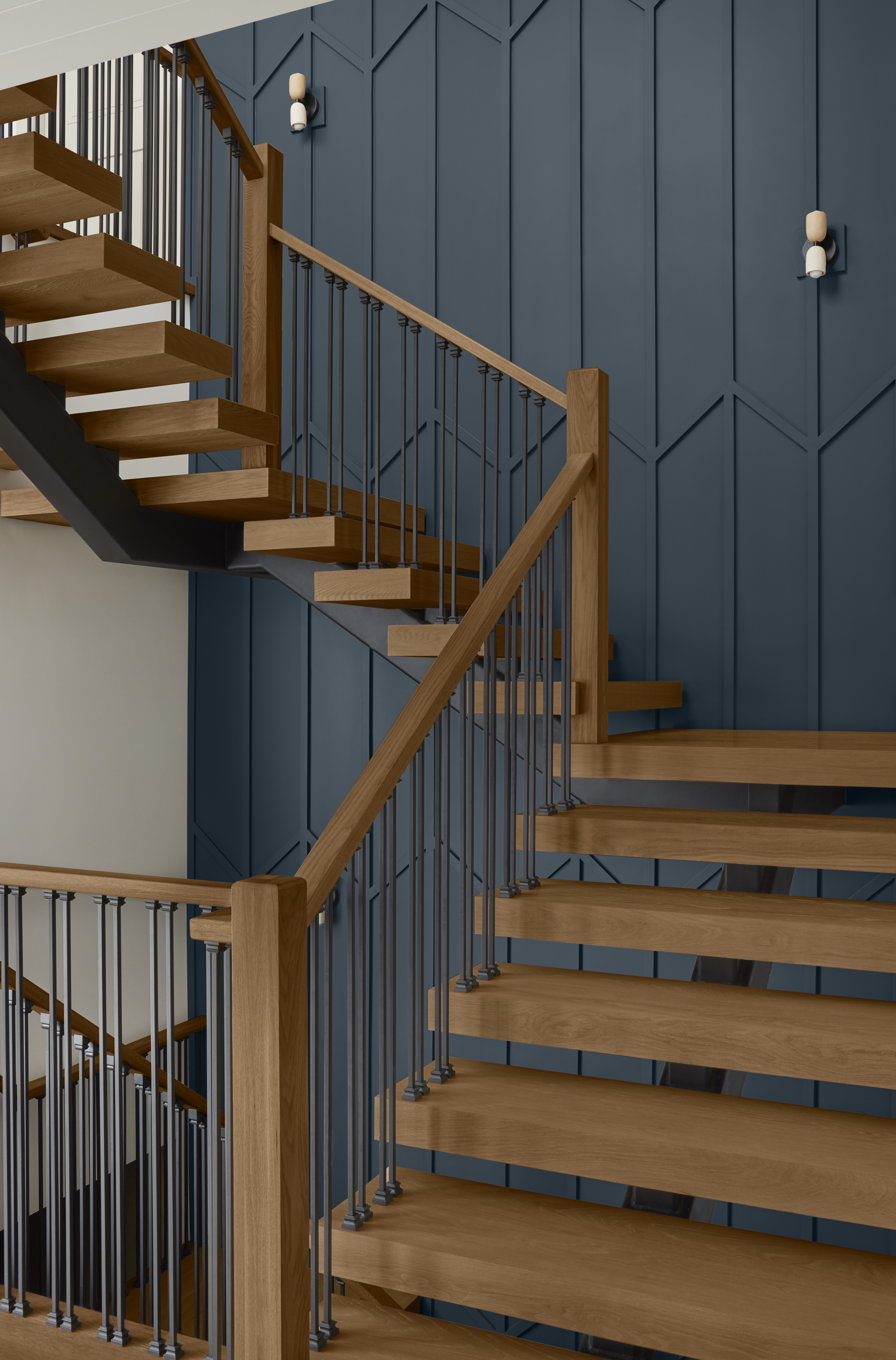 A modern staircase with wooden steps and a sleek combination of wooden and metal railings, set against a classic dark blue paneled wall adorned with two minimalist white sconce lights. 