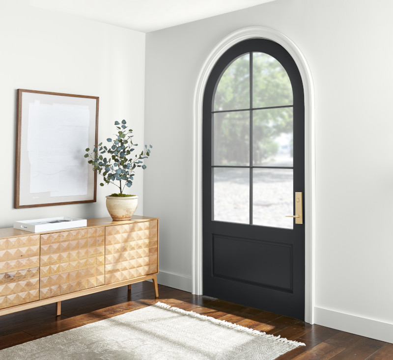 A home entrance with white walls and an arched black door. To the left, there is a wooden console table with patterned front panels, above which hangs a framed picture. The floor is covered by a beige area rug over hardwood flooring, and the space is well-lit by natural light coming through the glass part of the door. 