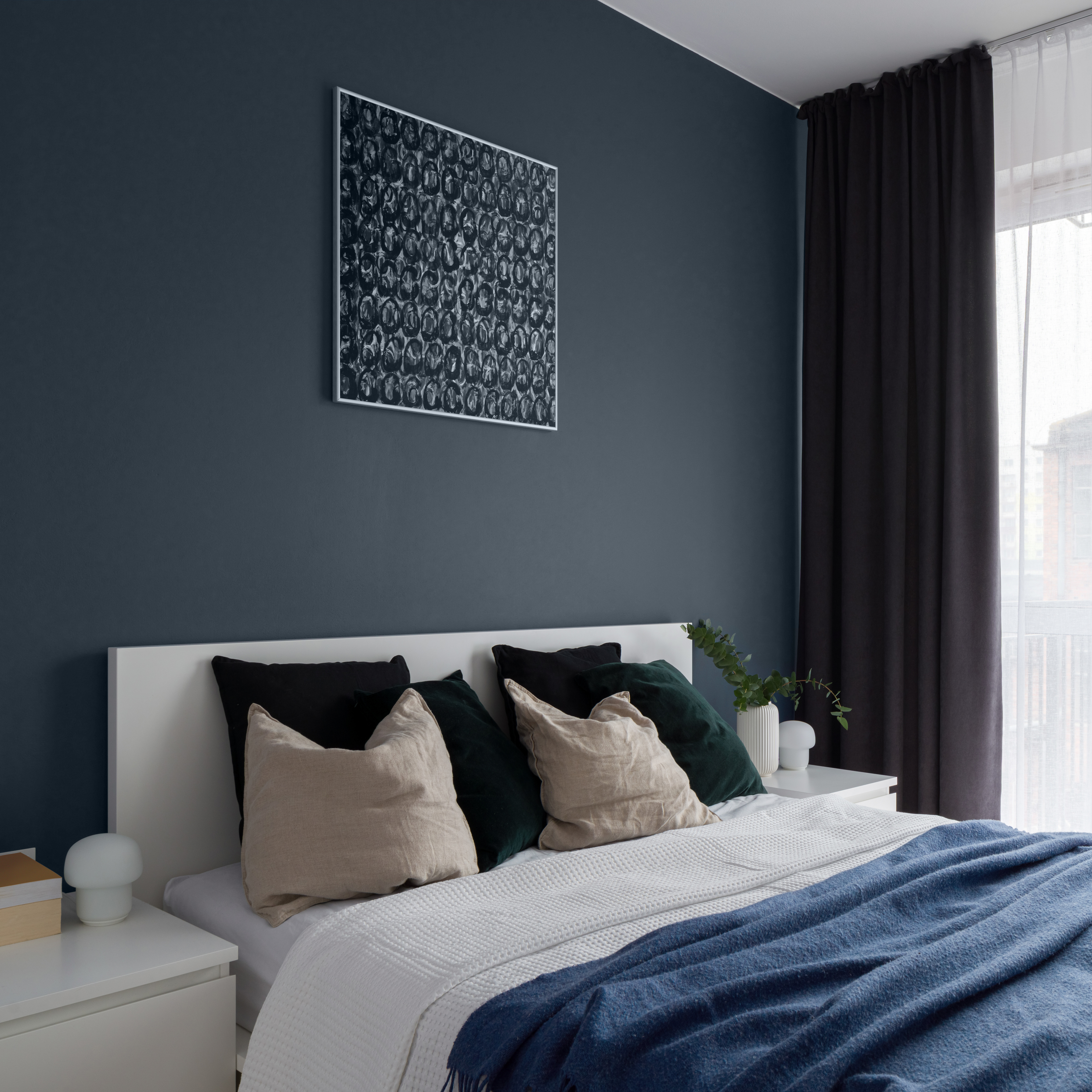 - A contemporary bedroom with a minimalist design, showcasing a white bed with crisp linens and a blue throw blanket. The room features a dark blue accent painted with a BEHR colour called Midnight Blue. The dark gray curtains are drawn back to let in natural light, complementing the serene and coordinated colour scheme of the room.