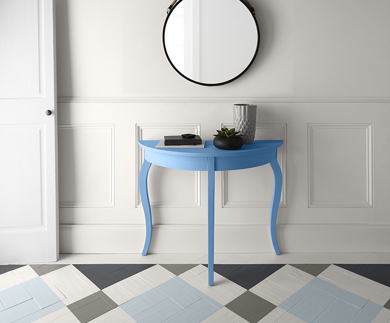 A hallway space featuring white walls, white trim, and a white door. A blue half-moon table is positioned against the wall beneath a round mirror with a black frame, adding a pop of colour to this setting. The floor has a geometric painted pattern of light and dark tiles, contributing to the room’s modern vibe.