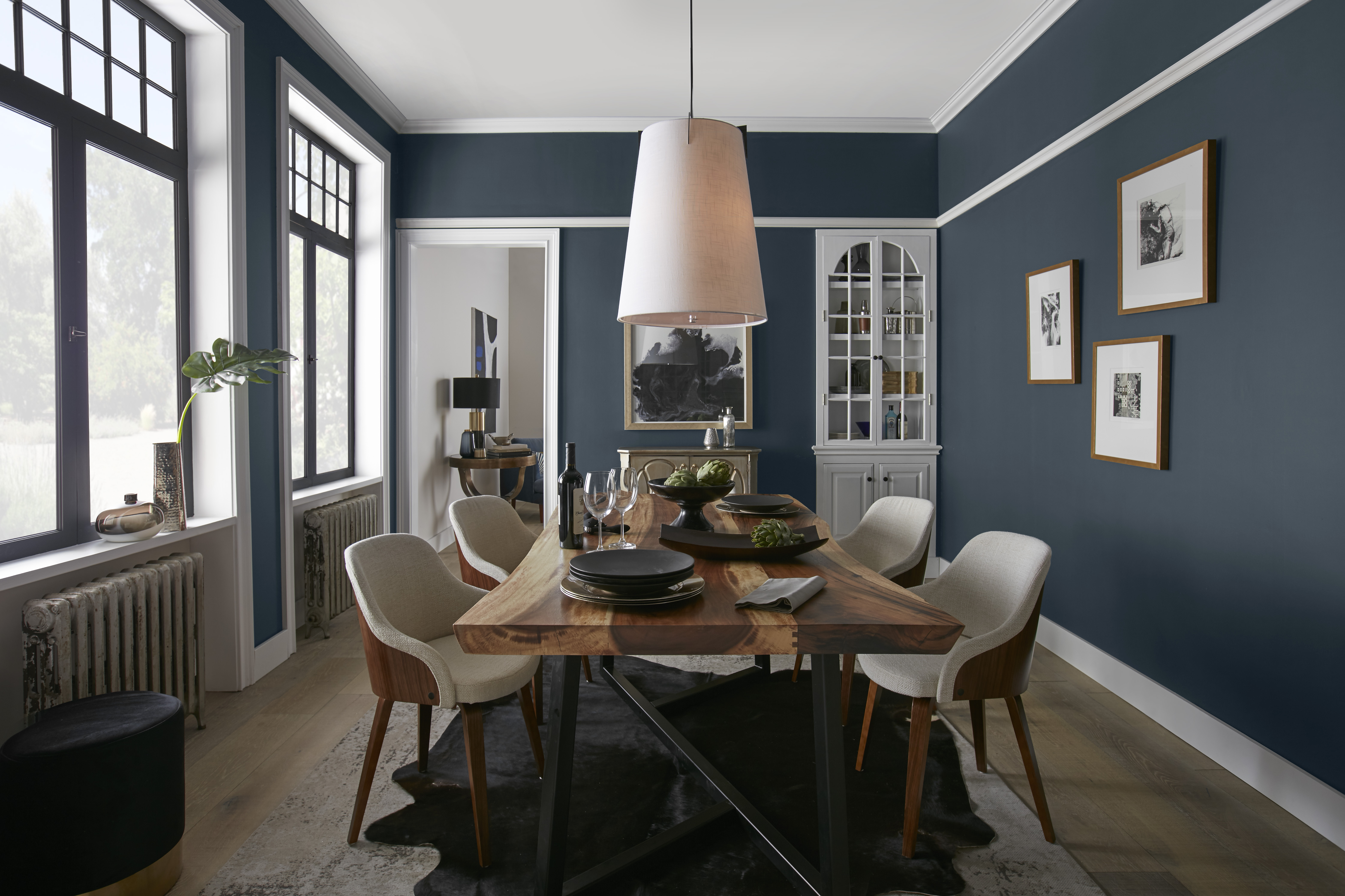 A chic dining space with a long wooden table surrounded by beige upholstered chairs, set against dark blue walls with white trim. A statement pendant light with a textured shade casts a soft glow on the table, which is neatly set with plates and glasses, and adorned with a green plant centerpiece. 