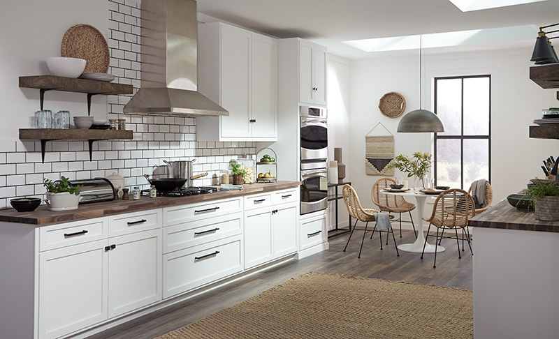 - A modern farmhouse kitchen featuring white walls and sleek white cabinetry with silver handles. The cabinets are complemented by a dark countertop and a white subway tile backsplash.  Open wooden shelves display various kitchen items. Stainless steel appliances, including a double oven, are built into the cabinetry.