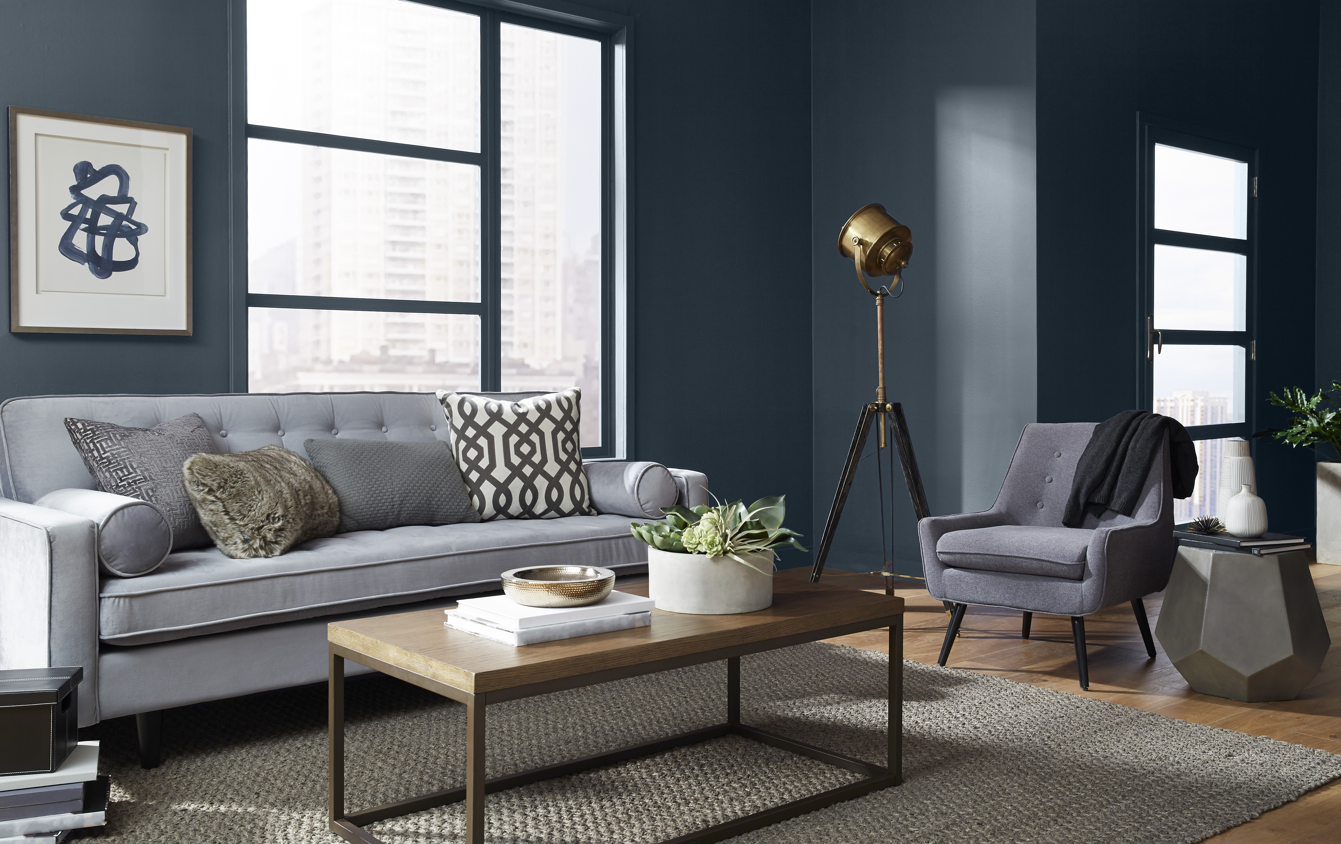 A cozy modern living room with a gray upholstered sofa, adorned with patterned throw pillows, and a matching armchair beside a unique geometric side table. A brass floor lamp adds warmth to the corner, while the deep blue walls, accented with white trim, frame large windows that reveal a cityscape view.