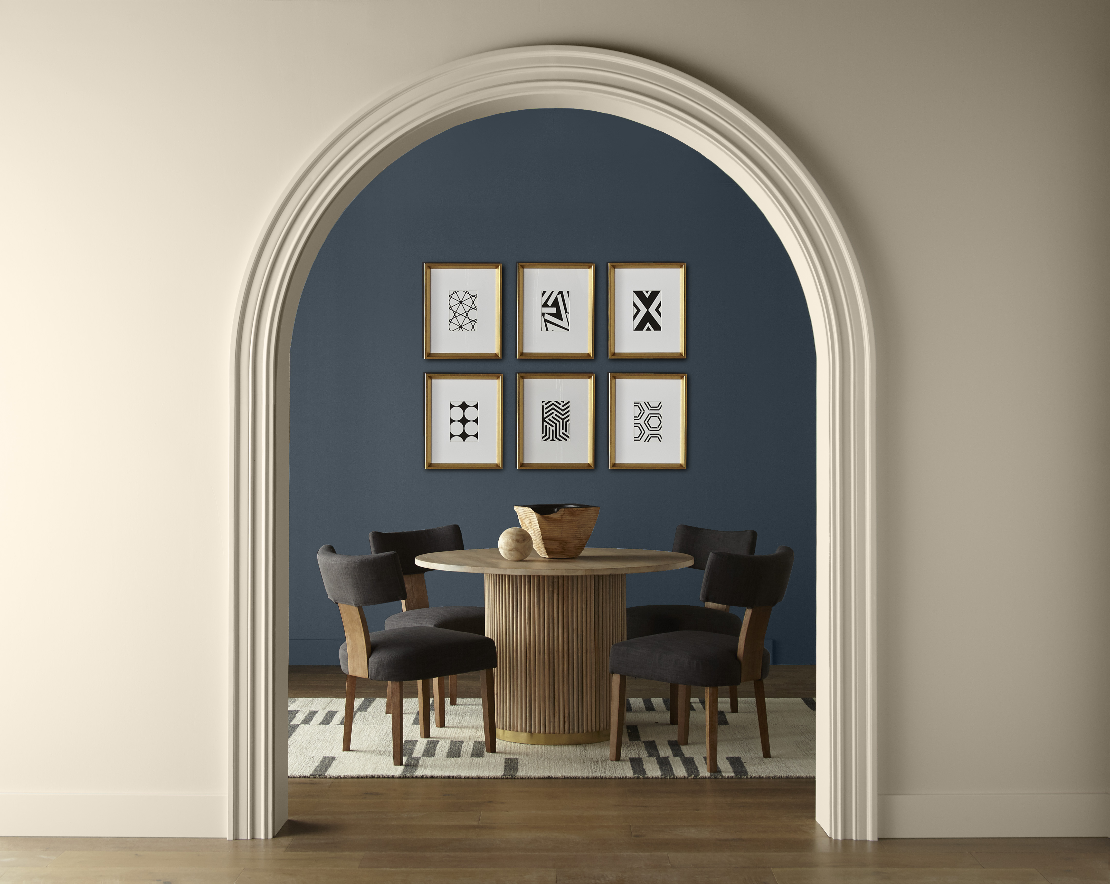 An elegantly designed dark blue dining area viewed through an arched doorway. The room features a round wooden table with four dark upholstered chairs around it. The floor is adorned with a light-coloured rug with darker geometric designs, complementing the artwork above. The foreground wall and trim are painted in a neutral tone, creating a warm and inviting atmosphere.