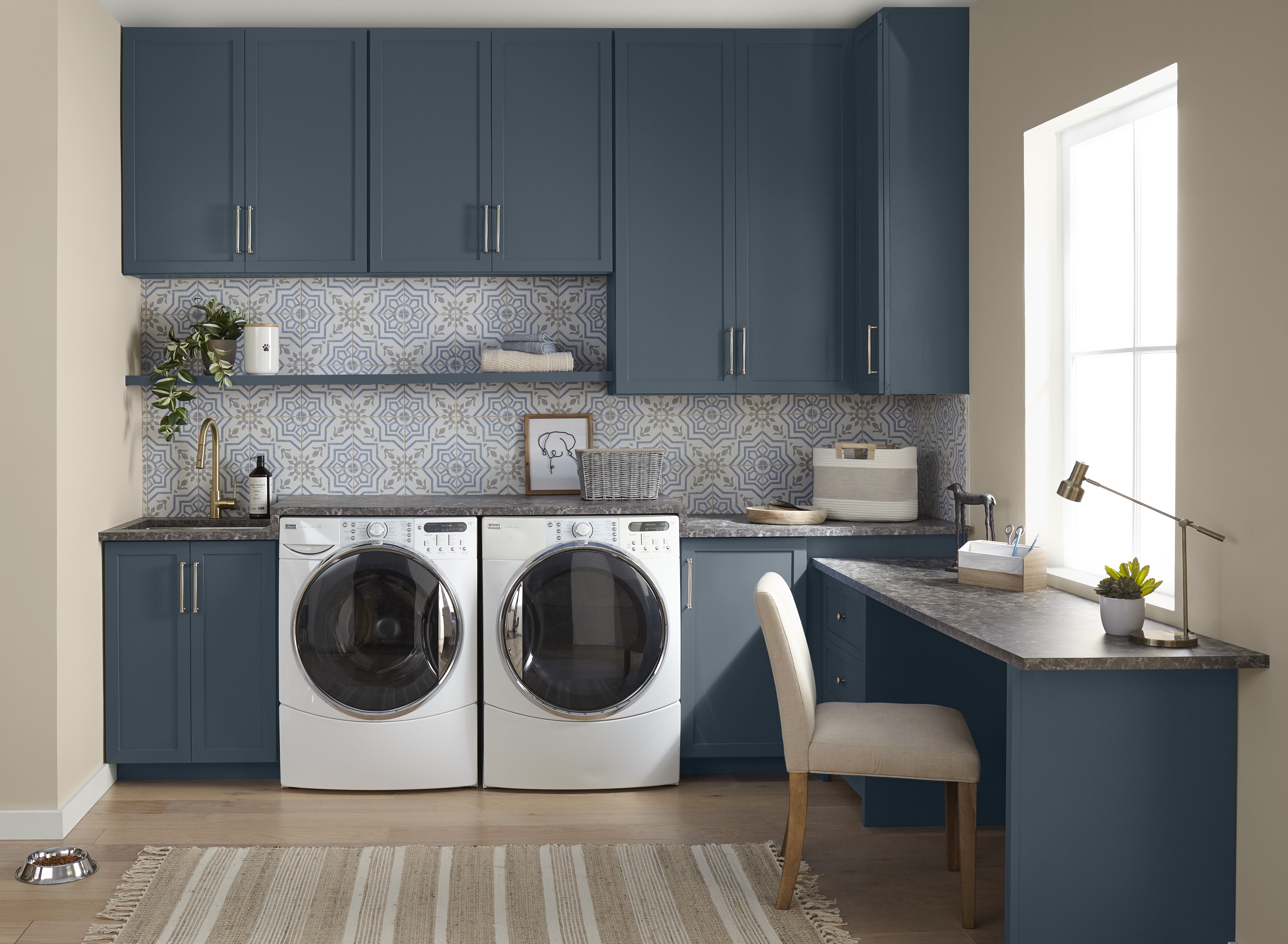 A laundry room-home office with dark blue cabinets that beautifully contrast with beige walls and a light grey and blue patterned tile backsplash. The trim is in Polar Bear, a bright white, adding to the fresh and clean look.