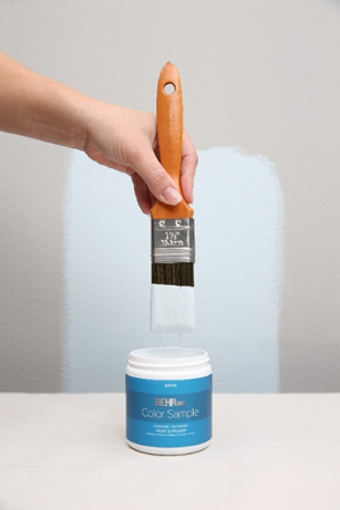 Image shows an 237 ml paint can open with a person holding a paint brush over it with paint dripping into the can. Wall behind has a swatch of painted colour.