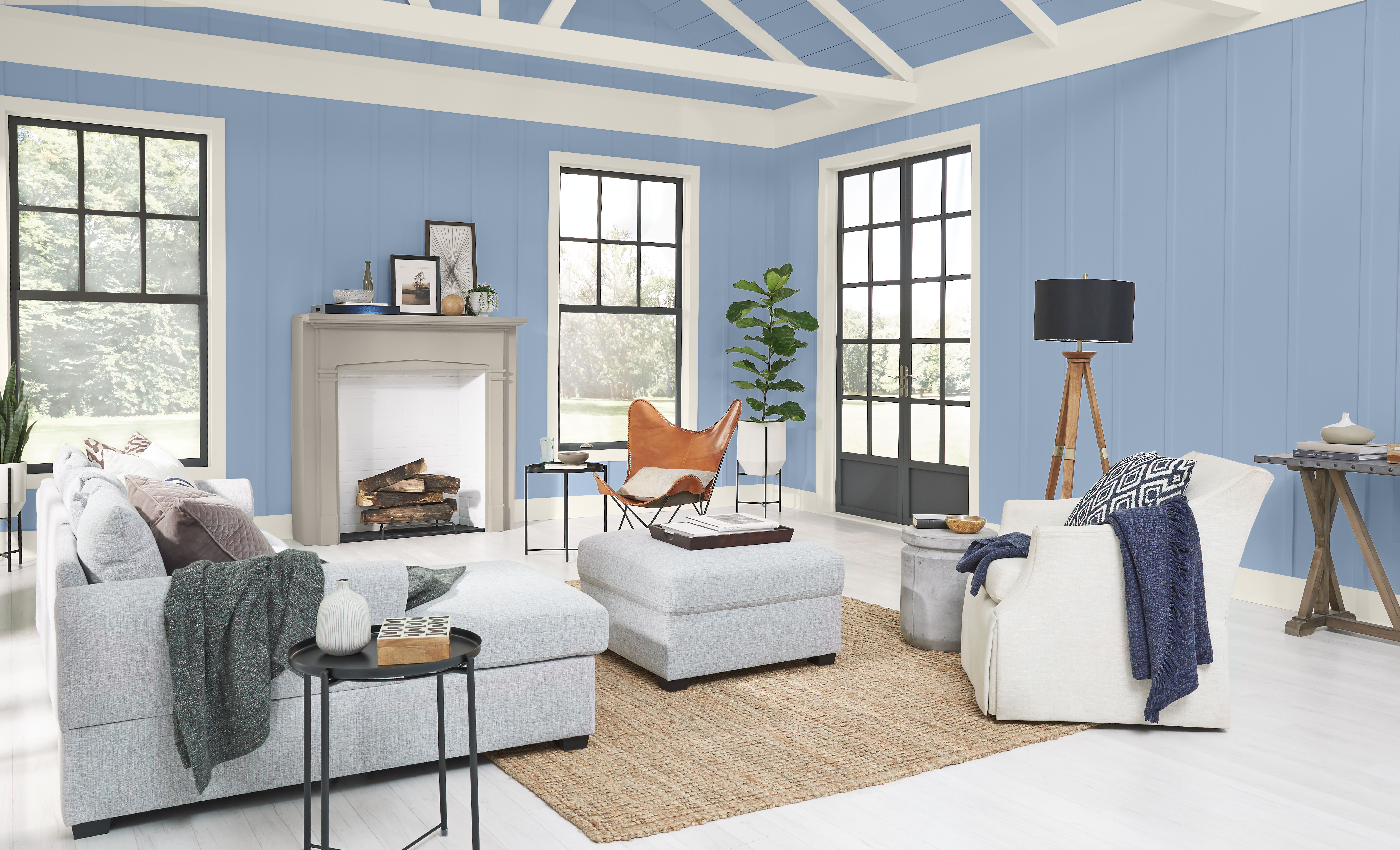 An airy living room featuring a grey fireplace mantle, blue walls, large black windows, and intricate ceiling woodwork. 